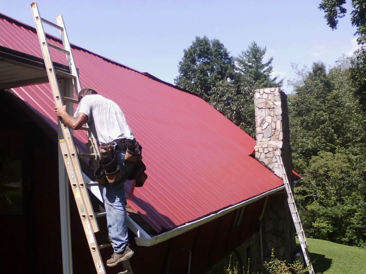 Roofer on a ladder beside a red metal roof and stone chimney — Soffit & Fascia Repair by Roofing Brothers in Turners Station